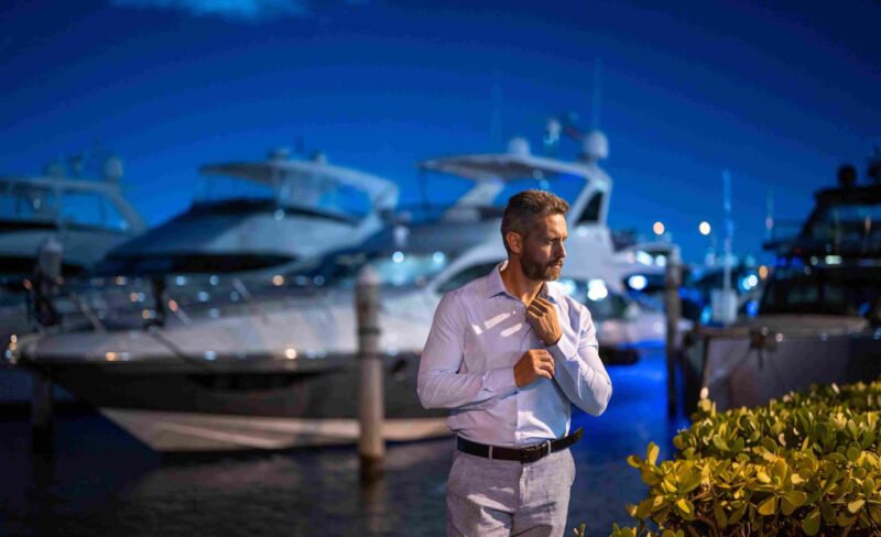 Well-dressed man standing near luxury yachts at a marina at night, representing affluence, waterfront living, and exclusive coastal communities.