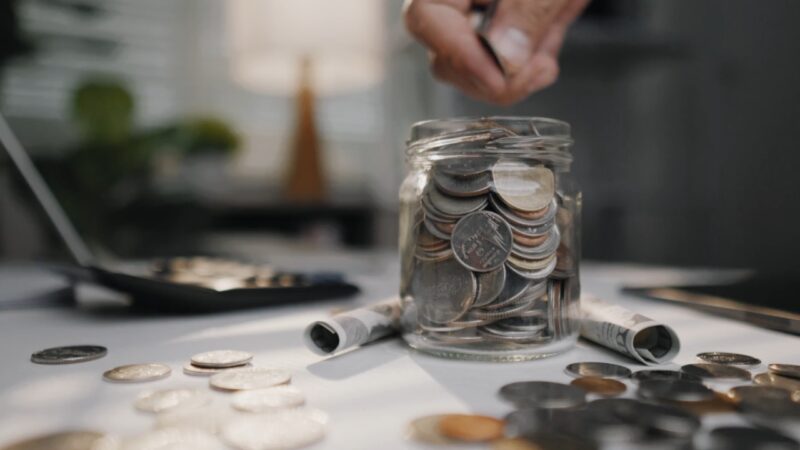 Hand drops coins into a jar on a desk with bills and loose change nearby